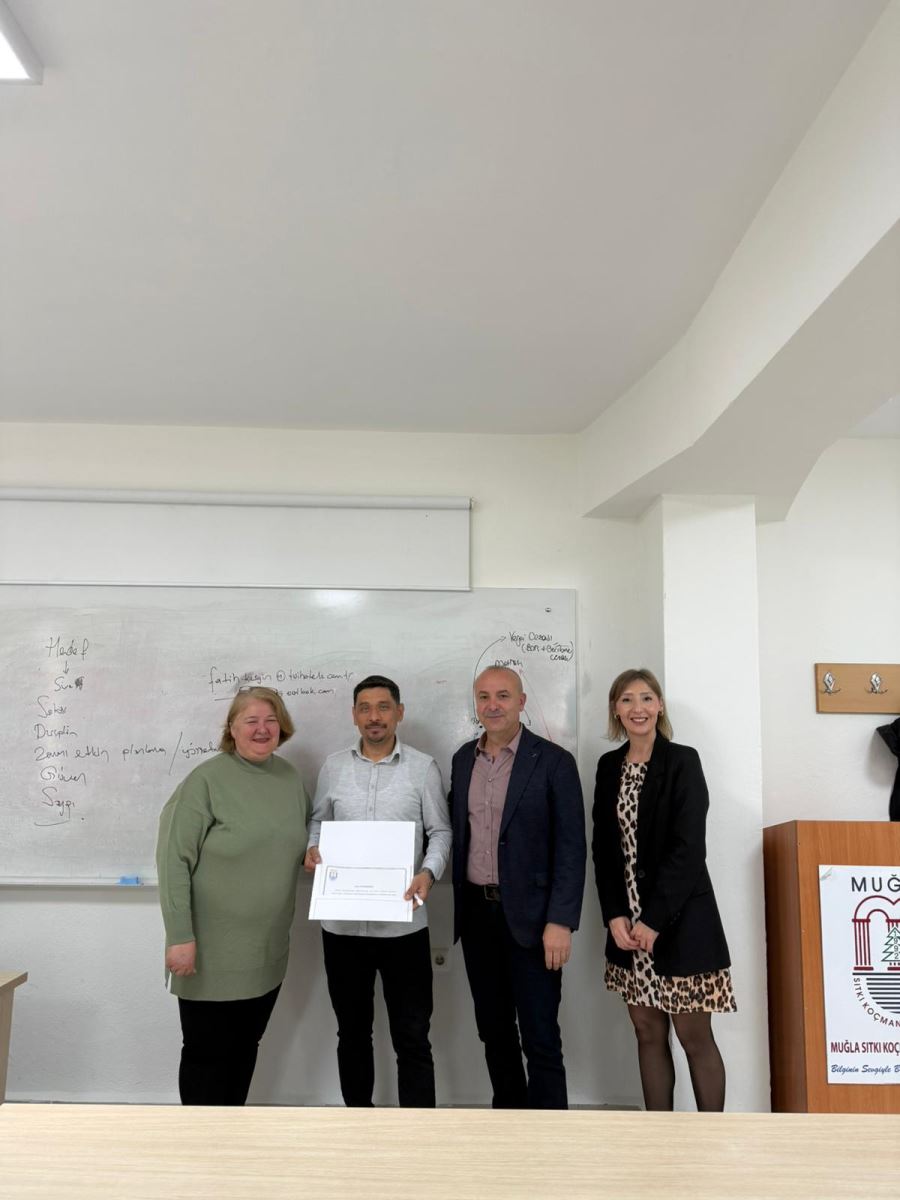 Bu fotoğraf, bir ödül töreninin daha geniş bir açıdan görüntüsünü sunuyor. Öğrenciler, öğretim üyeleri ve ödül alan kişi arasında bir etkileşim oluyor. Tahtada ders notları yazılı, ve arka planda ödül alan kişinin başı görünmüyor. Birkaç öğrenci, ellerinde telefonlarla bu anı izliyor.